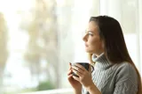 En person sidder med en kop kaffe og taenker, mens hun kigger ud af vinduet.