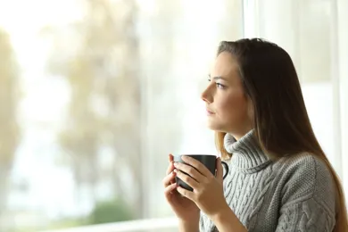 En person sidder med en kop kaffe og taenker, mens hun kigger ud af vinduet.