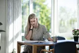 En kvinde sidder ved et bord, og holder sig på nakken, mens hun laeser en avis