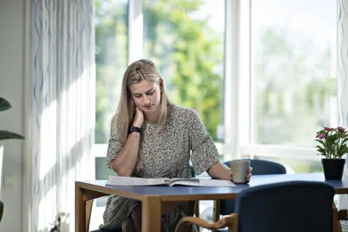En kvinde sidder ved et bord, og holder sig på nakken, mens hun laeser en avis